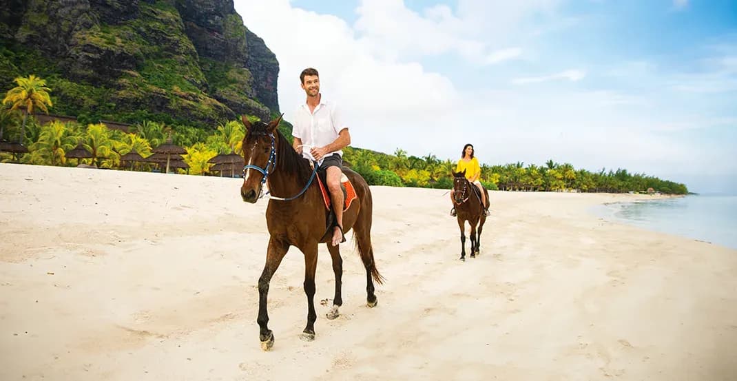 Guests horse riding along the white-sand beach at Dinarobin Beachcomber with Le Morne Brabant in the background