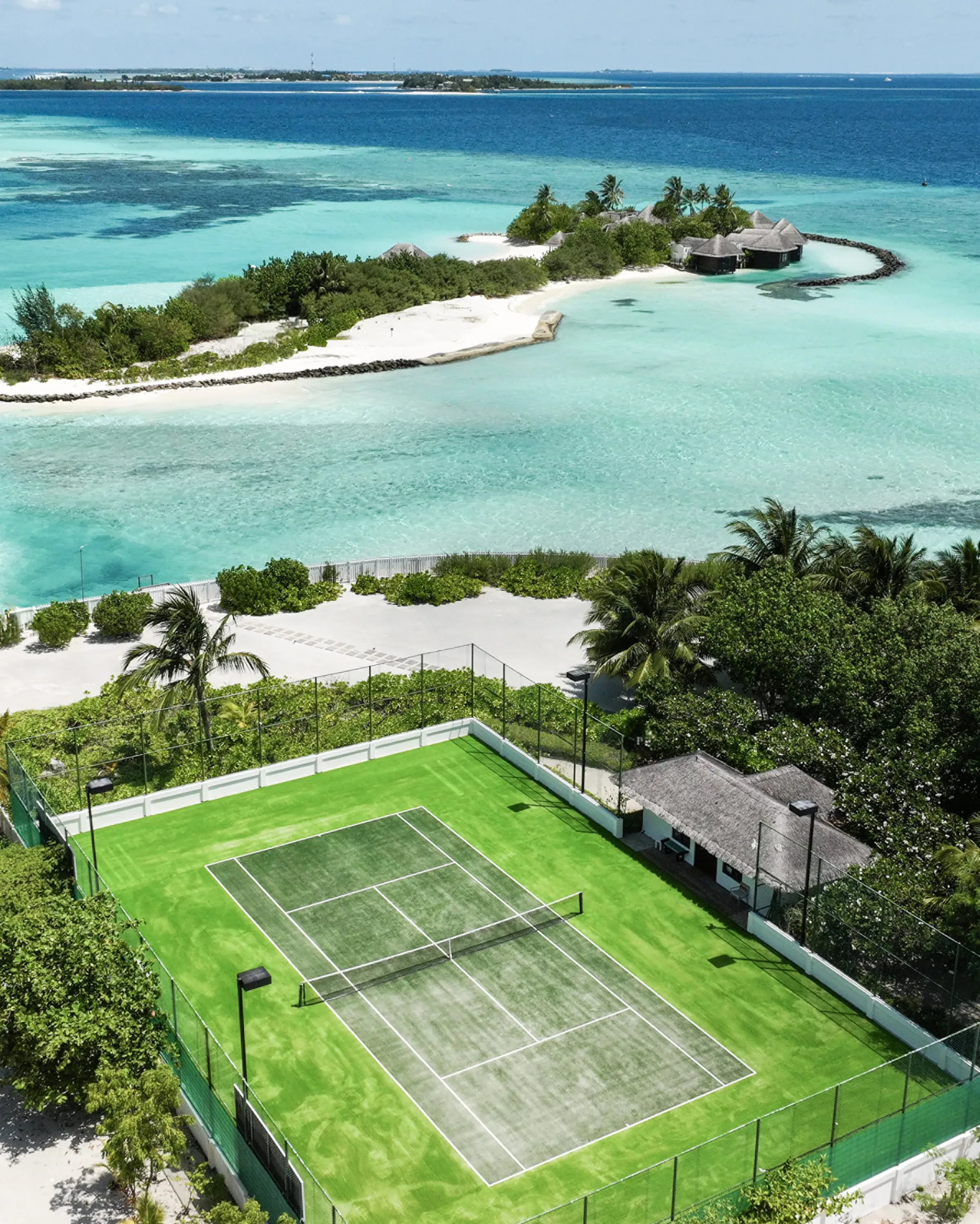 Aerial view of the tennis court and lagoon at Four Seasons Resort Maldives at Kuda Huraa, with the ŪRJĀ Naturopathy Island spa visible across the water