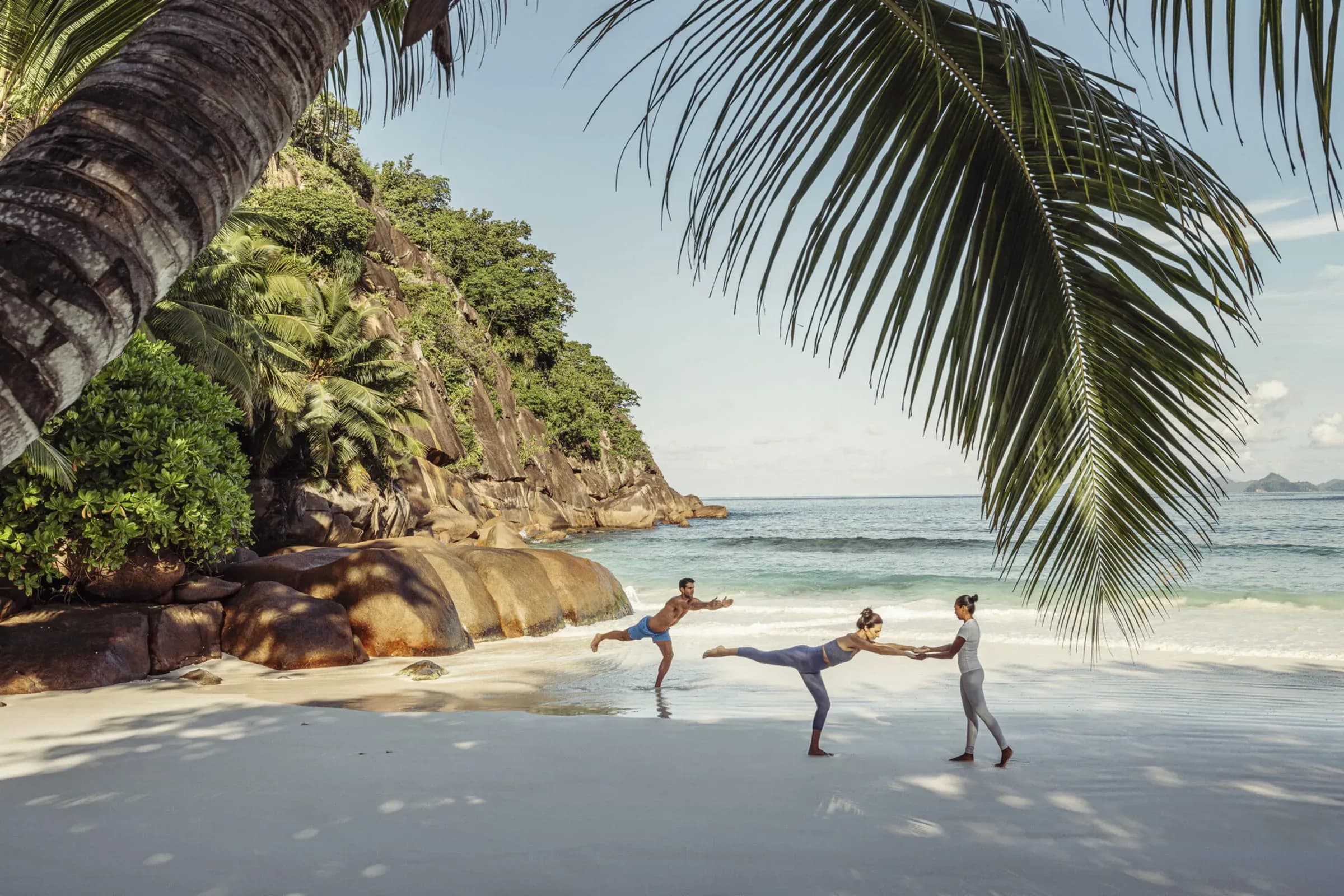 Guests and an instructor practising yoga on the beach beneath a large palm tree at Four Seasons Resort Seychelles