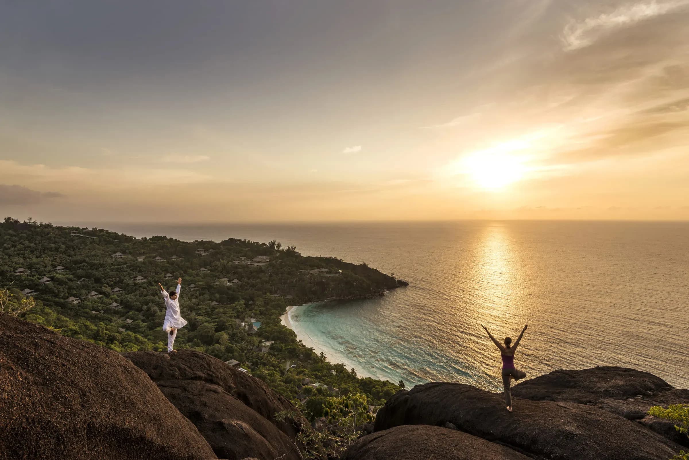 A couple stands on granite boulders above the tree canopy at Four Seasons Resort Seychelles, looking out to the ocean at sunrise