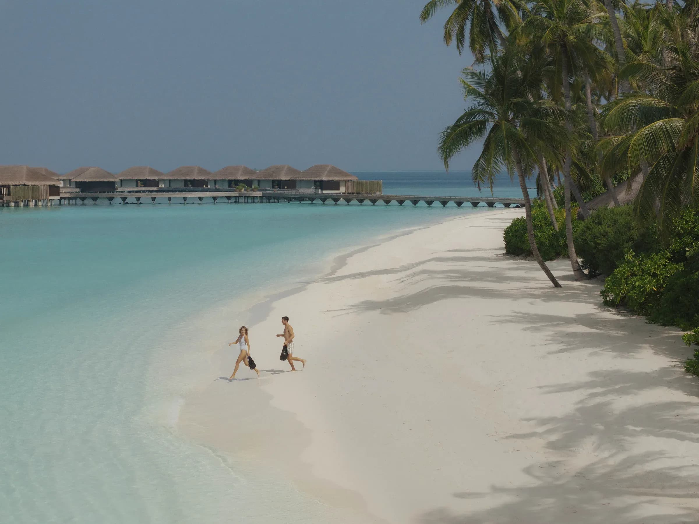Guests walking along the beach at Velaa Private Island with an overwater villa in the background